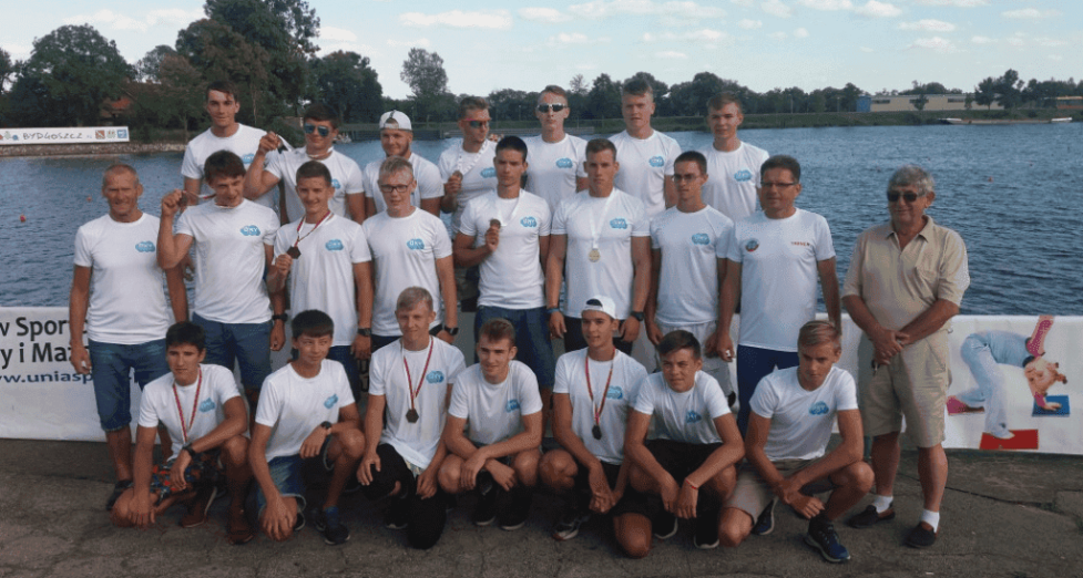 Zawodnicy i trenerzy na gali zwycięzców podczas zawodów w sportach wodnych na jeziorze, w tle woda i drzewa, promującym działalność OXynet Jarosław i sport wodny.